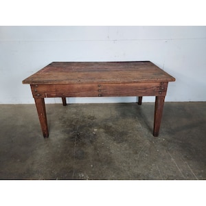 May include: A rustic wooden table with a rectangular top and four legs. The table is made of dark brown wood and has a distressed finish. The table is sitting on a concrete floor.