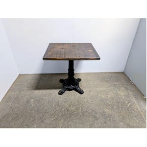 May include: A square table with a dark brown wooden top featuring a visible grain pattern. The table is supported by a single central pedestal base made of black metal, which branches out into four ornate, curved feet at the bottom.
