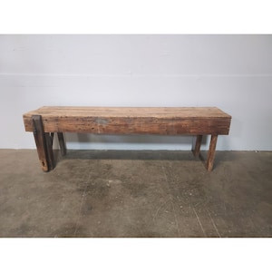 Puede incluir: Un banco de madera rústico con un diseño sencillo. El banco está hecho de tablas toscas y tiene un acabado de madera natural. Tiene una parte superior rectangular y dos pares de patas que se inclinan ligeramente hacia adentro. El banco mide aproximadamente 1,5 metros de largo y 45 centímetros de ancho.
