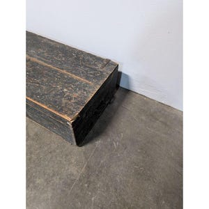May include: A rectangular, weathered wooden chest painted black. The chest has visible wood grain and is positioned at an angle against a white wall and a gray concrete floor. The chest appears to be vintage or antique.