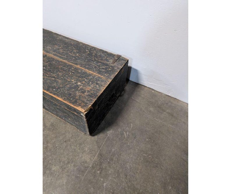 May include: A rectangular, weathered wooden chest painted black. The chest has visible wood grain and is positioned at an angle against a white wall and a gray concrete floor. The chest appears to be vintage or antique.