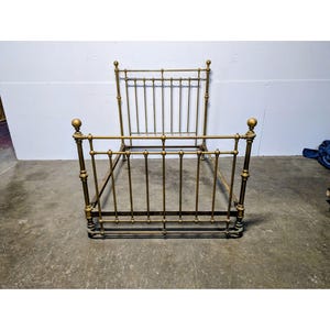 Op de afbeelding: Een bedframe van messing in vintage-stijl met een hoog hoofdeinde en een lager voeteneinde. Het frame is opgebouwd uit verticale metalen spijlen en is aan elk van de vier hoekstijlen voorzien van decoratieve bolvormige knoppen, wat zorgt voor een klassieke en symmetrische structuur.