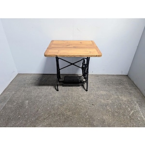 Puede incluir: Pequeña mesa cuadrada con una tapa de madera clara. La base es de metal negro, con un diseño intrincado que recuerda a una máquina de coser vintage. Una pieza de mobiliario única.