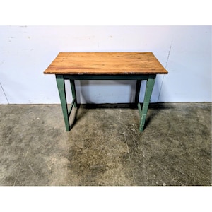 May include: A rectangular wooden table featuring a natural light brown wood grain top surface and four sturdy legs painted in a muted sage green colour. The table has a simple construction with a horizontal apron connecting the four legs.