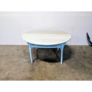 Puede incluir: Una mesa redonda con un tablero de madera de color claro y veta visible. La base está construida con mimbre pintado de azul claro, mostrando un diseño de celosía tejida alrededor del faldón y cuatro patas robustas ligeramente abiertas.