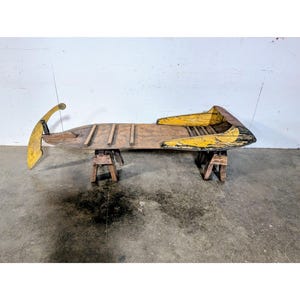 Op de afbeelding: Een antieke houten slee met gele verfaccenten. De slee wordt ondersteund door twee houten zaagbokken. Het ontwerp van de slee heeft een gebogen voorkant en een platte, plankvormige body, met tekenen van slijtage.