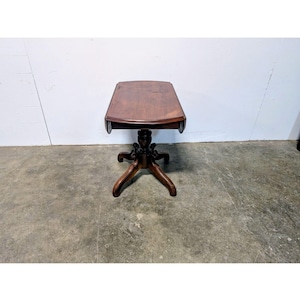 May include: A small, antique wooden table with a dark brown finish. The table has a rectangular top with rounded edges and a pedestal base with three curved legs. The table is approximately 24 inches tall.