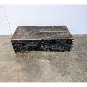 May include: A rectangular, weathered wooden trunk with a black finish and visible wood grain. The trunk has a hinged lid and metal hardware. Red paint is visible on the top. The trunk is on a concrete floor.