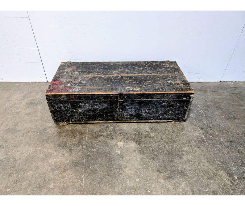 May include: A rectangular, weathered wooden trunk with a black finish and visible wood grain. The trunk has a hinged lid and metal hardware. Red paint is visible on the top. The trunk is on a concrete floor.