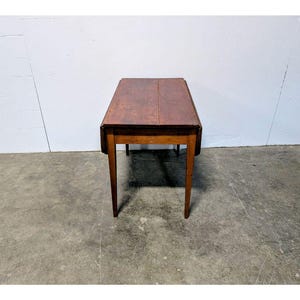 Puede incluir: Una pequeña mesa de madera antigua con una tapa rectangular y cuatro patas delgadas. La tapa es de un marrón oscuro intenso, y las patas son de un tono marrón más claro. La mesa mide aproximadamente 76 cm de largo.