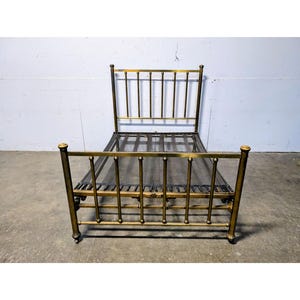 May include: A vintage brass bed frame with a headboard and footboard. The bed frame has vertical bars and rounded posts. The bed frame is made of metal and has a dark mesh base. The bed frame is on a concrete floor.