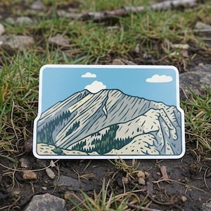 May include: A rectangular sticker with a mountain range illustration. The design includes a blue sky with clouds, a light blue background, and a detailed depiction of a mountain with trees. The sticker is placed on a grassy surface with rocks.