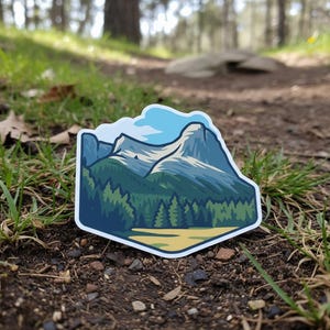 May include: A die-cut sticker depicting a mountain scene with a forest and a yellow field. The sticker has a white border and is placed on a dirt path with grass.