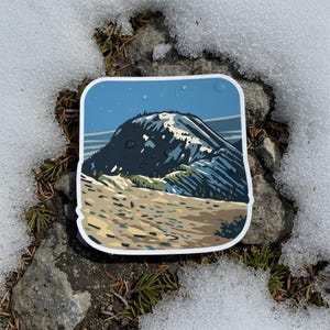 May include: A square sticker depicting a mountain landscape with a blue sky and a snow-covered peak. The sticker has a white border and is placed on a surface of snow and rocks, with scattered pine needles.
