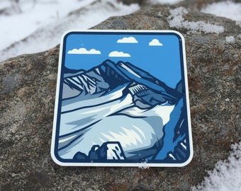 May include: A square sticker featuring a mountain range scene. The design includes a blue sky with white clouds, snow-covered peaks, and a valley. The sticker is placed on a gray rock with snow.