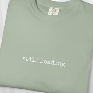 May include: Sage green t-shirt with the words "still loading" in white, centered on the front. The shirt has a crew neck and a tag visible at the neckline. The fabric appears soft and comfortable.