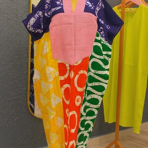 K&ouml;nnte beinhalten: Ein locker sitzendes Kaftan-Kleid mit Color-Blocking-Design und einem einfarbigen rosa V-Ausschnitt-Einsatz. Das Kleidungsst&uuml;ck zeigt Abschnitte aus blauem, gelbem, orangefarbenem und gr&uuml;nem Stoff, die jeweils mit wei&szlig;en Batik-Mustern verziert sind.