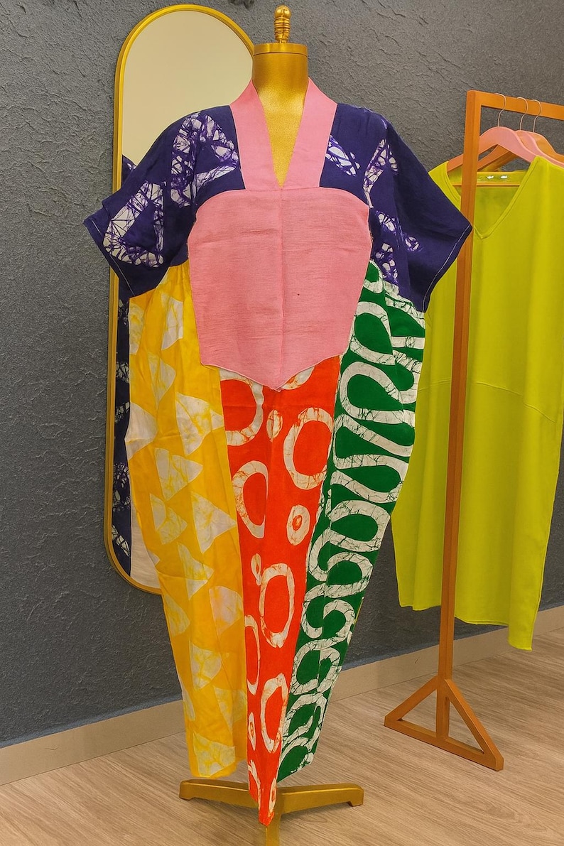 K&ouml;nnte beinhalten: Ein locker sitzendes Kaftan-Kleid mit Color-Blocking-Design und einem einfarbigen rosa V-Ausschnitt-Einsatz. Das Kleidungsst&uuml;ck zeigt Abschnitte aus blauem, gelbem, orangefarbenem und gr&uuml;nem Stoff, die jeweils mit wei&szlig;en Batik-Mustern verziert sind.