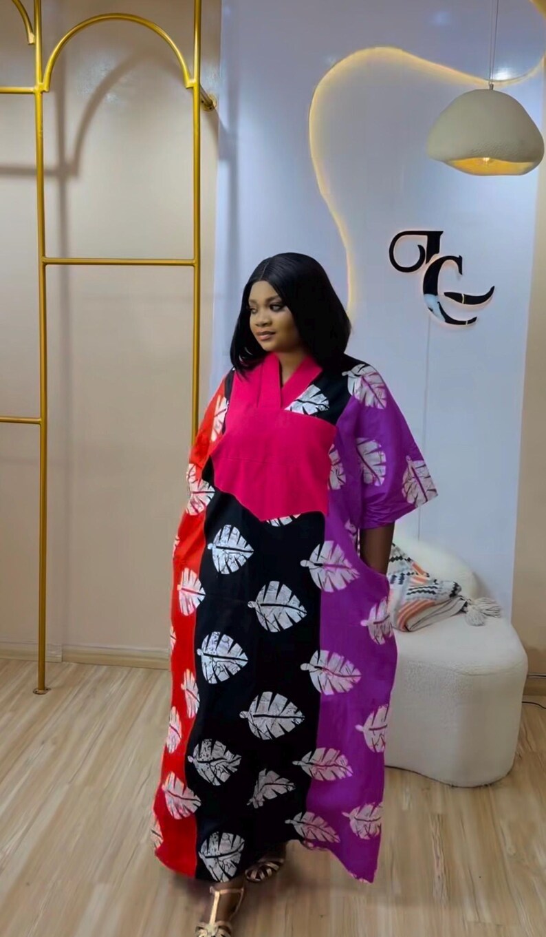 K&ouml;nnte beinhalten: Ein locker sitzendes Kaftan-Kleid mit einem lebendigen Color-Block-Design in Rot, Schwarz und Lila. Das Kleidungsst&uuml;ck hat einen V-Ausschnitt, kurze &Auml;rmel und ein sich wiederholendes wei&szlig;es Blattmuster auf den schwarzen und lila Stoffteilen.