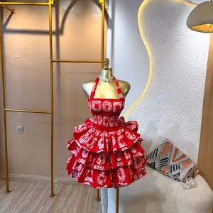 Puede incluir: Un vestido halter rojo y blanco con estampado, con un corpiño fruncido y una falda de varios niveles con volantes. La tela muestra un motivo circular repetitivo en toda la prenda, que se exhibe en un torso de maniquí de color dorado.