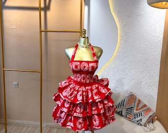 Könnte beinhalten: Ein rot-weiß gemustertes Neckholder-Kleid mit gesmoktem Oberteil und einem mehrlagigen, gerüschten Rock. Der Stoff zeigt ein sich wiederholendes kreisförmiges Motiv auf dem gesamten Kleidungsstück, das auf einem goldenen Schaufensterpuppen-Torso präsentiert wird.