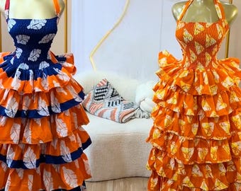 Könnte beinhalten: Zwei ärmellose Kleider auf Schaufensterpuppen. Das linke Kleid hat ein blaues Oberteil mit weißen Blattmustern und orangefarbenen Stufenrüschen. Das rechte Kleid ist komplett orange mit weißen Blattmustern, Neckholder und vielen Rüschen.