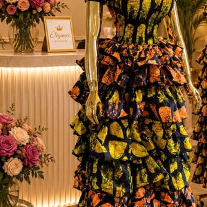 Peut inclure: Une robe longue jusqu'au sol avec des manches courtes bouffantes et une encolure carrée, présentant une base bleu marine foncé avec un imprimé géométrique abstrait jaune et orange vif. Le corsage est smocké et la jupe comporte des volants.