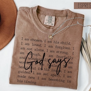 May include: Espresso-colored t-shirt with the text "God says" in a script font. The shirt features a list of affirmations in a simple, sans-serif font. A silver necklace with a pendant is draped across the shirt.
