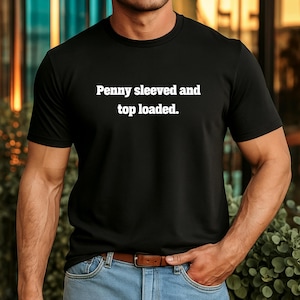 Könnte beinhalten: Schwarzes T-Shirt mit Rundhalsausschnitt und dem weißen Text "Penny sleeved and top loaded." Das Shirt ist aus weichem Material und hat kurze Ärmel. Das Model trägt eine blaue Jeans und einen braunen Gürtel.