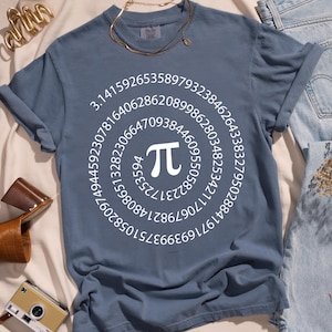 Puede incluir: Una camiseta azul polvorienta con un símbolo Pi blanco y un patrón circular de números. La camiseta está combinada con mulas de cuero marrón, un collar dorado y una cámara vintage. La camiseta está hecha de un material suave.