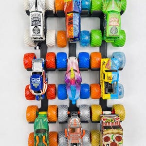 Pode incluir: Um suporte de exibição preto montado na parede com nove monster trucks de brinquedo. Os caminhões são de várias cores, incluindo verde, laranja, azul e amarelo, com gráficos detalhados e pneus grandes. O texto "Holds 9 Monster Trucks" está no topo.