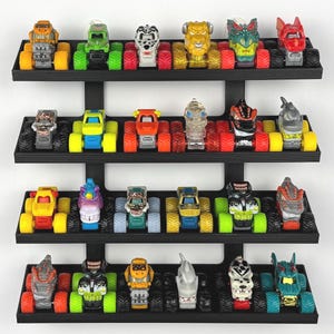May include: A collection of toy monster trucks displayed on a black tiered shelf. The trucks feature various colors and designs, including monster faces and themed graphics. The tires are large and colorful.