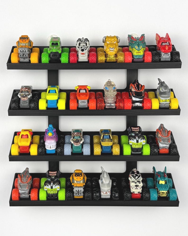 May include: A collection of toy monster trucks displayed on a black tiered shelf. The trucks feature various colors and designs, including monster faces and themed graphics. The tires are large and colorful.