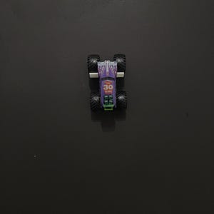 Puede incluir: Un peque&ntilde;o cami&oacute;n monstruo de juguete morado y blanco con neum&aacute;ticos negros grandes. El cami&oacute;n tiene el texto "30 YEARS". El cami&oacute;n est&aacute; sobre una superficie oscura.