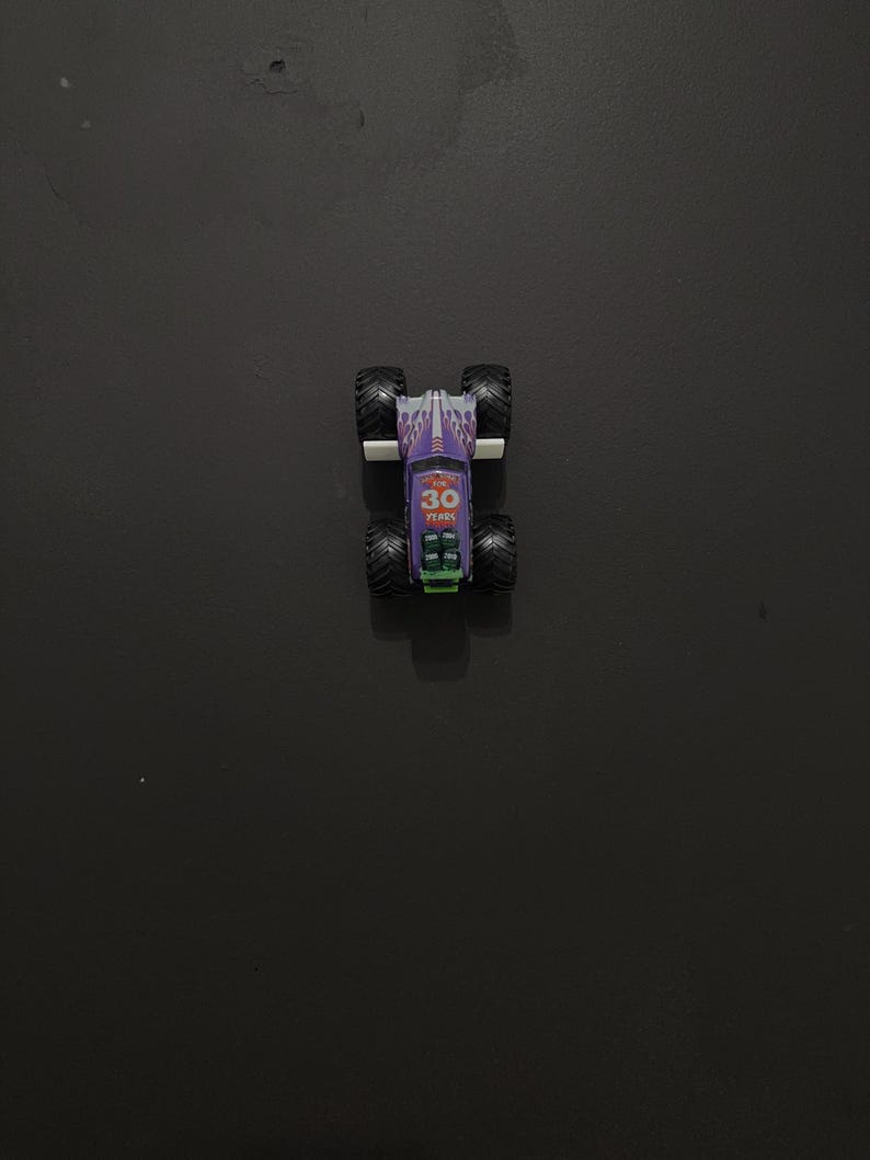 Puede incluir: Un peque&ntilde;o cami&oacute;n monstruo de juguete morado y blanco con neum&aacute;ticos negros grandes. El cami&oacute;n tiene el texto "30 YEARS". El cami&oacute;n est&aacute; sobre una superficie oscura.