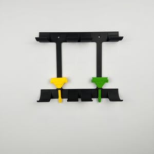 May include: Black wall-mounted tool holder with two colored tools. The holder has multiple slots and a rectangular frame. One tool is yellow, the other is green, both with text. The background is white.