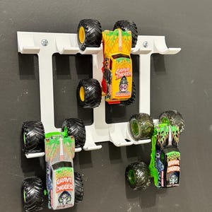 Puede incluir: Un organizador de camiones de juguete blanco montado en la pared que contiene varios monster trucks de juguete. Los camiones son de varios colores, incluyendo verde, amarillo y plateado, con neum&aacute;ticos negros grandes. Los camiones tienen la inscripci&oacute;n "Grave Digger".
