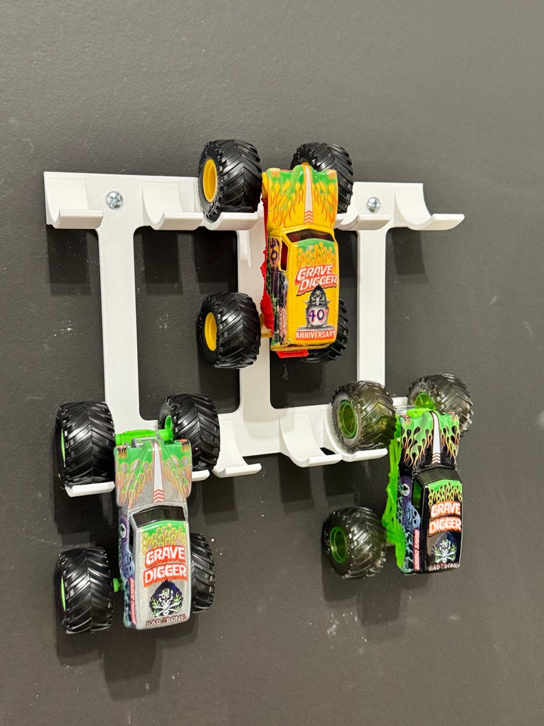Puede incluir: Un organizador de camiones de juguete blanco montado en la pared que contiene varios monster trucks de juguete. Los camiones son de varios colores, incluyendo verde, amarillo y plateado, con neum&aacute;ticos negros grandes. Los camiones tienen la inscripci&oacute;n "Grave Digger".