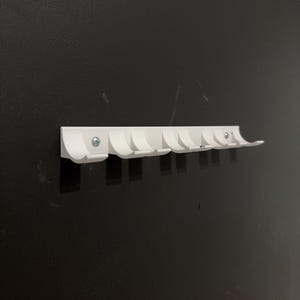 Puede incluir: Un perchero blanco montado en la pared con ocho ganchos curvos. El perchero est&aacute; hecho de un material s&oacute;lido y se fija a una pared oscura con tornillos. Los ganchos est&aacute;n espaciados uniformemente y dise&ntilde;ados para colgar abrigos, sombreros o bolsos. Mide unos 30 cm de largo.