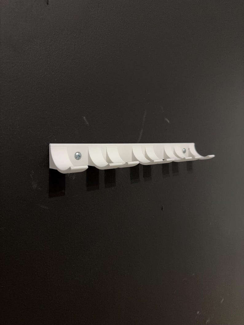 Puede incluir: Un perchero blanco montado en la pared con ocho ganchos curvos. El perchero est&aacute; hecho de un material s&oacute;lido y se fija a una pared oscura con tornillos. Los ganchos est&aacute;n espaciados uniformemente y dise&ntilde;ados para colgar abrigos, sombreros o bolsos. Mide unos 30 cm de largo.