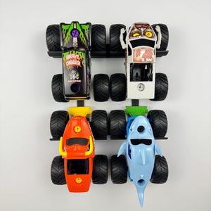 May include: Four toy monster trucks in a row, each with large black tires. The trucks are in various colors, including black with green flames, white with a monster face, orange, and light blue shaped like a shark.