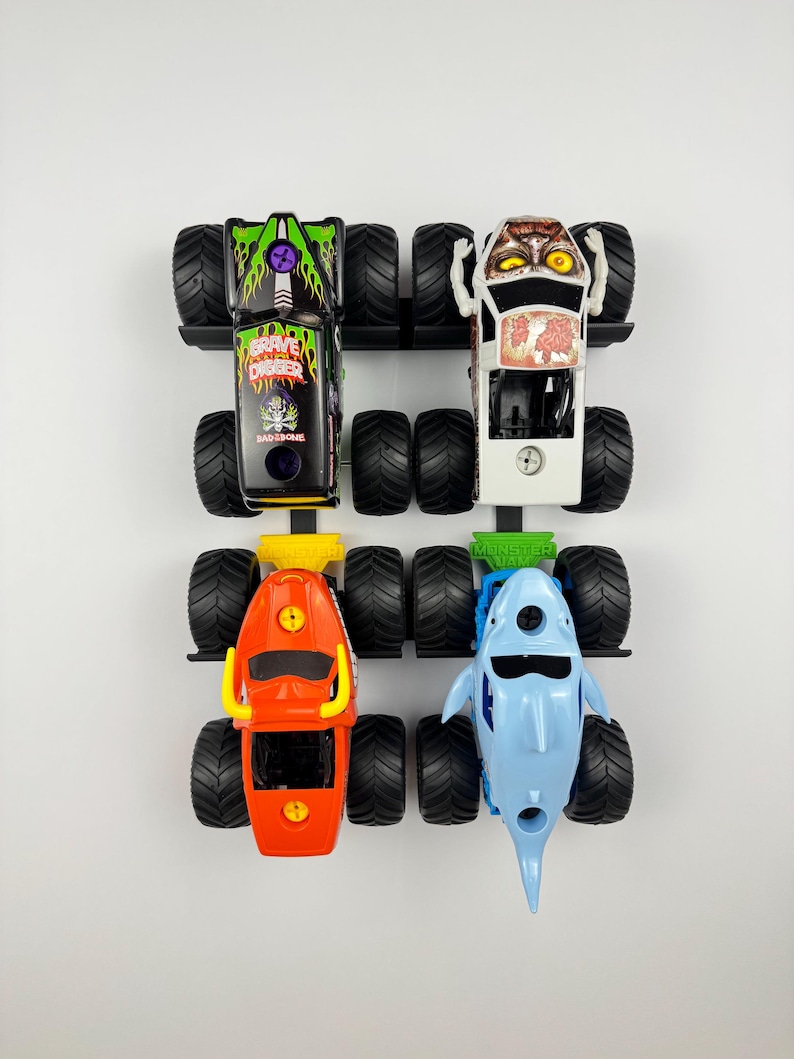 May include: Four toy monster trucks in a row, each with large black tires. The trucks are in various colors, including black with green flames, white with a monster face, orange, and light blue shaped like a shark.