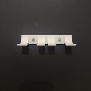 Puede incluir: Un colgador de pared de pl&aacute;stico blanco con cuatro ganchos curvos. El soporte rectangular est&aacute; fijado a la pared con dos tornillos plateados. Los ganchos est&aacute;n espaciados uniformemente y sobresalen hacia abajo.