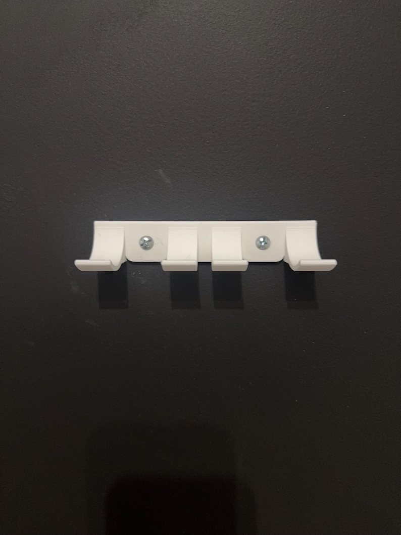 Puede incluir: Un colgador de pared de pl&aacute;stico blanco con cuatro ganchos curvos. El soporte rectangular est&aacute; fijado a la pared con dos tornillos plateados. Los ganchos est&aacute;n espaciados uniformemente y sobresalen hacia abajo.