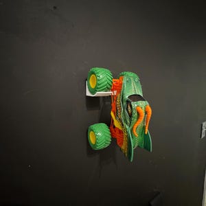 May include: A green and orange monster truck toy with large green tires and yellow rims. The truck has a detailed design with orange tentacles and a green body. The toy is mounted on a white wall mount against a black wall.