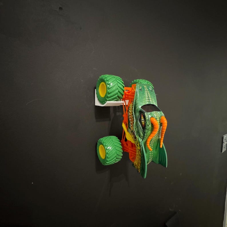 May include: A green and orange monster truck toy with large green tires and yellow rims. The truck has a detailed design with orange tentacles and a green body. The toy is mounted on a white wall mount against a black wall.