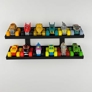 May include: A collection of colorful toy monster trucks displayed on two black shelves. The trucks feature various designs, including a shark, a monster, and a skull. The shelves are mounted on a white wall.