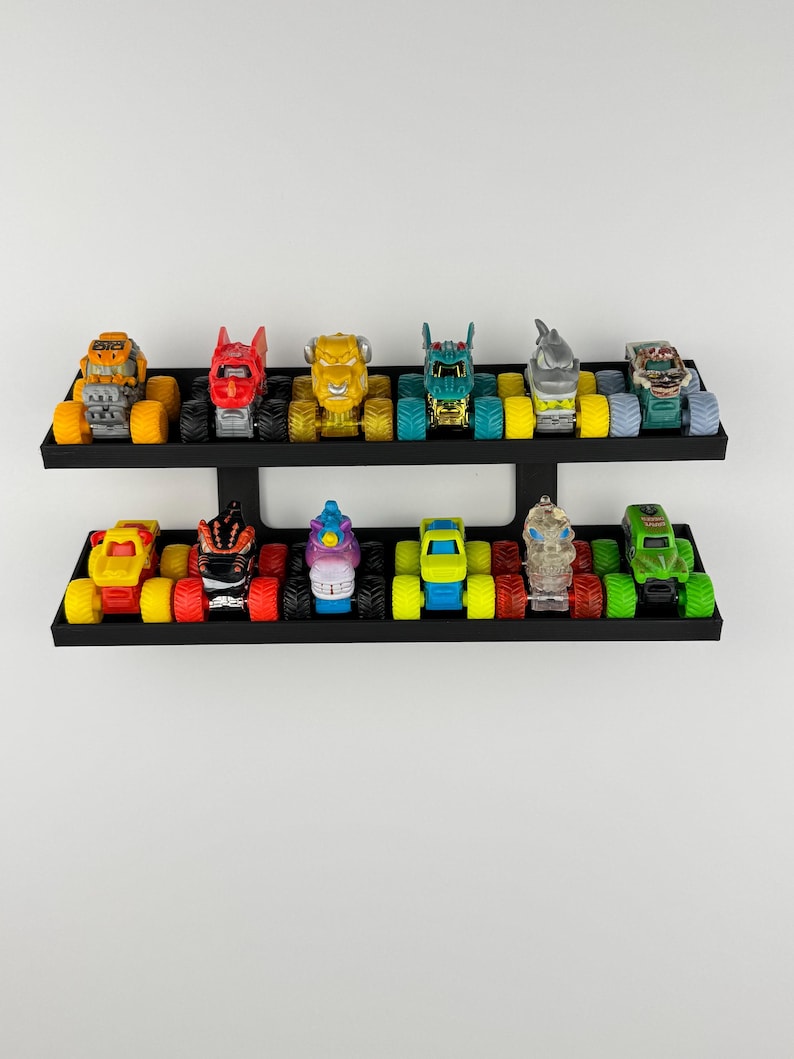 May include: A collection of colorful toy monster trucks displayed on two black shelves. The trucks feature various designs, including a shark, a monster, and a skull. The shelves are mounted on a white wall.