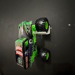 May include: A green, black, and white Grave Digger monster truck toy. The truck features large black tires with green rims and the words "Grave Digger" and "Bad Bone" on the side. Flames and skull graphics decorate the body.