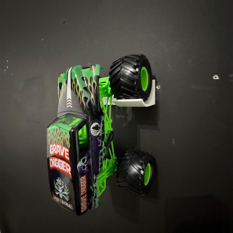 May include: A green, black, and white Grave Digger monster truck toy. The truck features large black tires with green rims and the words "Grave Digger" and "Bad Bone" on the side. Flames and skull graphics decorate the body.